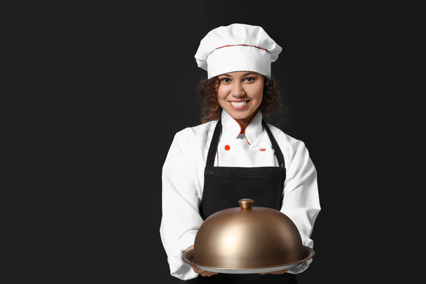Female African-American chef with tray and cloche on dark background - 写真・画像