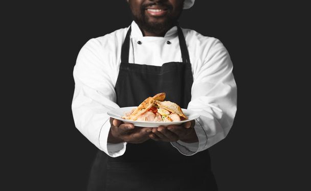 Male African-American chef with tasty dish on dark background - Foto, Imagen