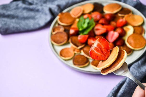 Assiette avec crêpes traditionnelles et minuscules crêpes aux fraises et feuilles de menthe sur fond sombre. Nourriture tendance. Concentration sélective sur la fourchette avec de minuscules crêpes et fraises - Photo, image