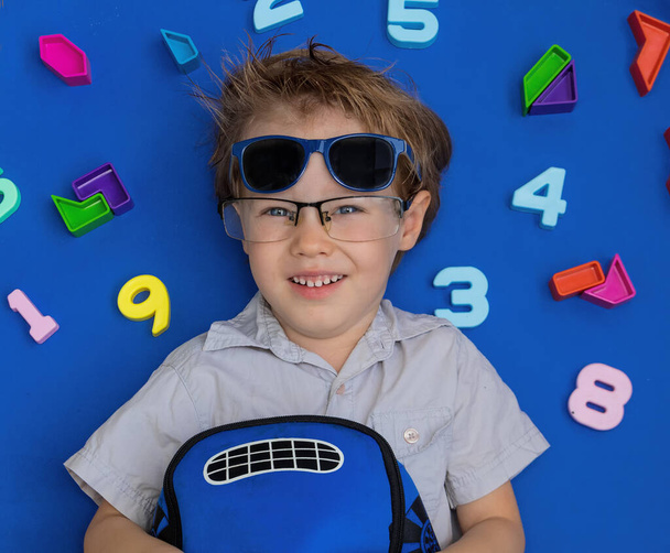 back to school. Kid boy took off his sunglasses and put on his glasses for sight. First-grader kid is with a backpack on a blue background among the numbers - Fotó, kép