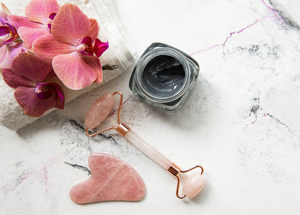 Skincare routine di bellezza mattutina nel bagno di casa massaggiatore rullo di giada viso per la terapia cosmetica benessere, maschera e fiori di orchidea rosa su sfondo di marmo - Foto, immagini