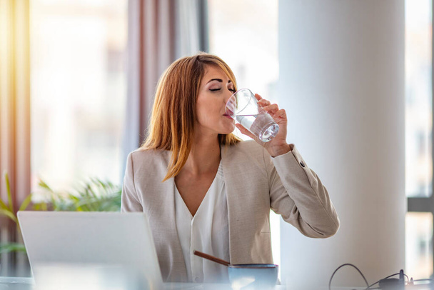 Office worker drinking water working on line. Young businesswoman drinking water in office. Happy beautiful young woman drinking water. Smiling female model holding transparent glass in her hand. - Фото, зображення