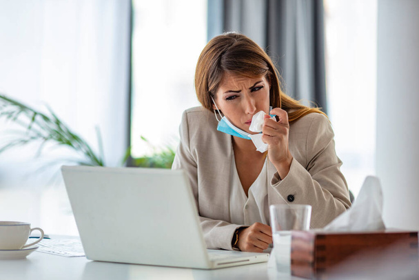 Cropped shot of an attractive young businesswoman sitting alone in her office and coughing as she suffers from a cold. Medical mask and hand disinfectant and stressed woman. Cropped shot of a businesswoman working in her office while suffering from a - Фото, зображення