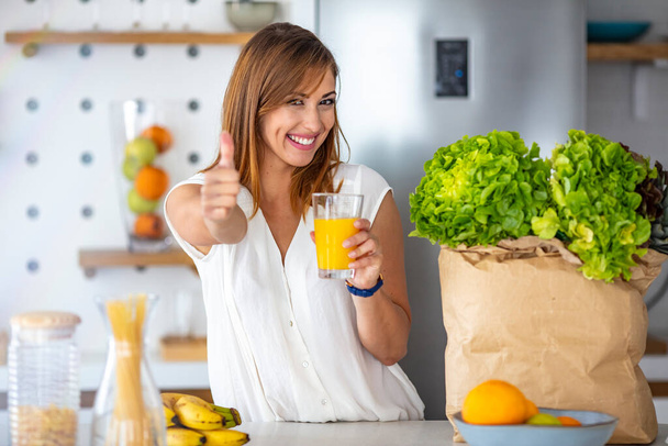 Young woman looking at camera while drinking orange juice in the kitchen at home. Woman with healthy food indoors. Morning routine. Young woman enjoying fresh orange juice - Foto, imagen