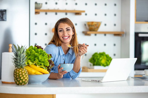 Beautiful young woman working with laptop while eating in the kitchen. Charming young female surfing internet at home. Always online. Woman surfing the net while enjoying breakfast in her kitchen at home - Foto, Imagem