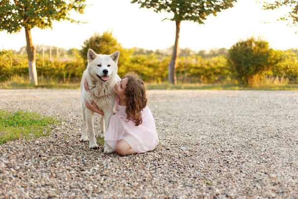 Nettes kleines Mädchen mit einem großen weißen Hund im Park. Ein schönes 7-jähriges Mädchen in rosa Kleid umarmt ihren Lieblingshund bei einem Sommerspaziergang. - Foto, Bild