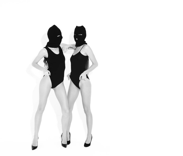 Two beautiful sexy women in black swimwear bathing suit. Models wearing bandit balaclava mask.Hot girls posing near white wall in studio.Seductive female in nice lingerie.Crime and violence - Photo, Image