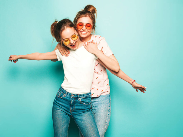 Dos jóvenes hermosas y rubias chicas hipster sonrientes en ropa de vaqueros hipster de verano de moda. Sexy mujeres despreocupadas posando cerca de la pared azul. Modelos modernos y positivos que se divierten en gafas de sol - Foto, imagen