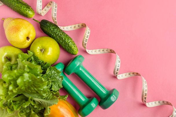 Összetétel súlyzókkal, zöld és sárga zöldségekkel és gyümölcsökkel, mérőszalag rózsaszín háttérrel, felülnézet. Az étrendi táplálkozás és az egészséges életmód fogalma. - Fotó, kép