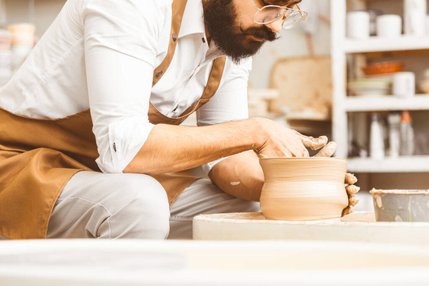 A young male potter is engaged in craft in his workshop on a potters wheel and makes a clay product - Fotografie, Obrázek