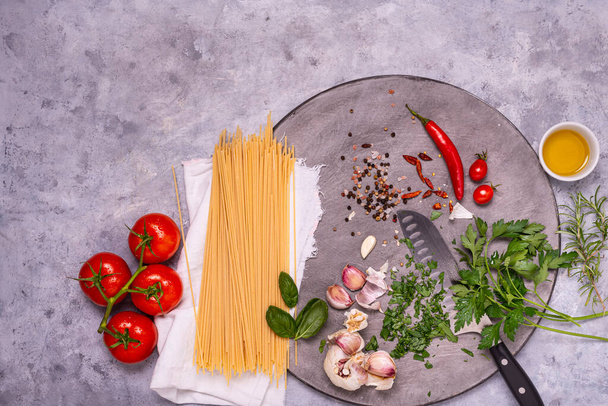 Italienische Küche: auf einem grauen Holzschneidebrett, rohe Spaghetti, Knoblauch, Olivenöl, Pfeffer und zahlreiche Zutaten und Kräuter, Draufsicht - Foto, Bild