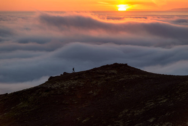 Silhueta de um homem no cume acima do mar de nuvens, montanhas nebulosas ao pôr do sol na Islândia
 - Foto, Imagem