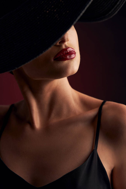 portrait of a beautiful tanned girl with professional makeup, red lips, on a burgundy background in a black dress with straps and black hat that covers her eyes - Fotó, kép