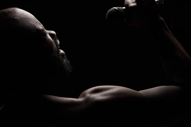 portret van een donkere huid gespierde knappe man met een baard op een zwarte achtergrond die emotioneel zingt in een microfoon - Foto, afbeelding