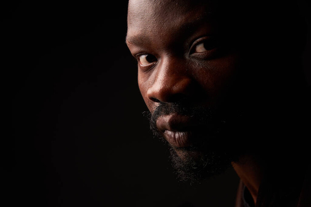 portrait photo of a dark-skinned handsome guy with a beard looking at the camera with serious face on a black background - Zdjęcie, obraz
