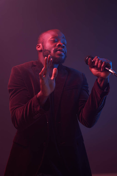 portrét pohledného muže tmavé pleti v tmavé bundě a tričku drží v rukou mikrofon a emocionálně zpívá v tmavém ateliéru s červeným a modrým světlem - Fotografie, Obrázek