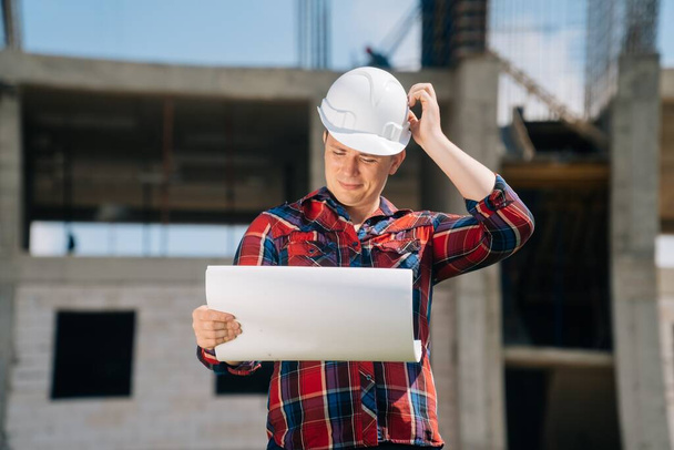 Portrait of man in awhite protective helmet with paper in his hands on a ckground of a building construction. The concept of a smart engineer on the background of a crane with blue sky of nature. - Fotó, kép