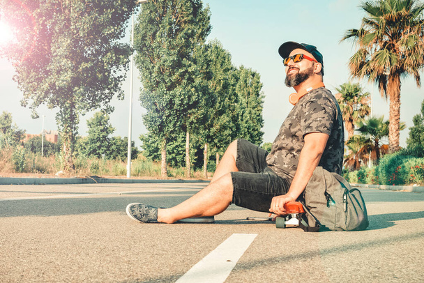 Gebogen man in korte broek zittend op een skateboard glimlachend. - Foto, afbeelding
