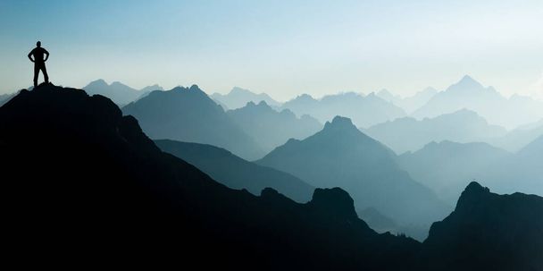 Hombre llegando a la cima después de escalar y caminar disfrutando de la libertad y mirando hacia las siluetas de las montañas panorama durante el amanecer
. - Foto, imagen