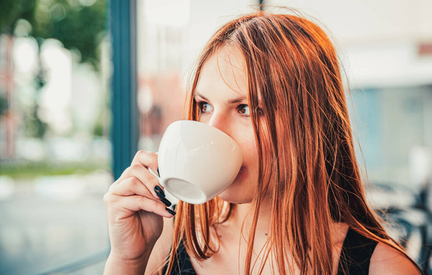 portrait of young teenager redhead girl with long hair with cup of hot coffee at city street cafe terrace - Fotoğraf, Görsel