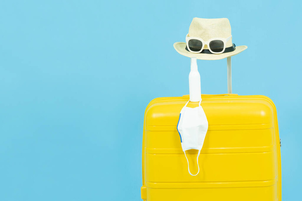 Summer holidays,vacation and travel concept.Yellow Suitcase or luggage bag,sun hat,sunglasses,medical face mask and alcohol Spray,the reopening of the Tourism after the quarantine on blue background - 写真・画像