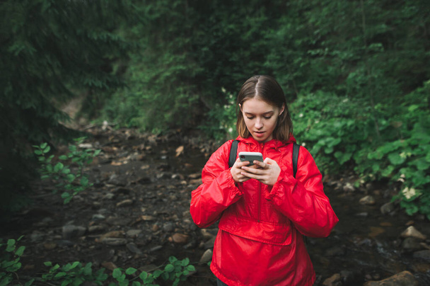 Young female hiker is concentrated on her smartphone, texting and feeling puzzled in the fir woods. Tourist girl in a red raincoat is googling the maps in the mountain forest. River on the background. - Photo, Image