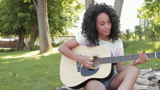 Uśmiechnięta Afroamerykanka grająca na gitarze akustycznej w parku - Materiał filmowy, wideo