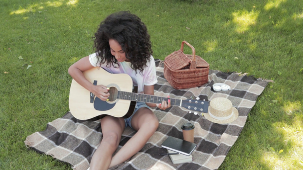 Χαμογελώντας αφροαμερικάνικη κοπέλα παίζει ακουστική κιθάρα κατά τη διάρκεια πικ-νικ στο πάρκο  - Πλάνα, βίντεο