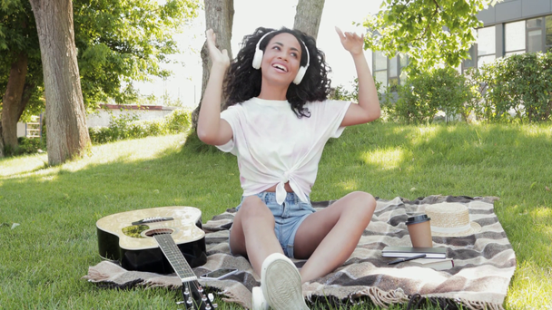 Χαμογελώντας αφροαμερικανή κοπέλα με ακουστικά χορεύοντας κατά τη διάρκεια πικνίκ στο πάρκο  - Πλάνα, βίντεο