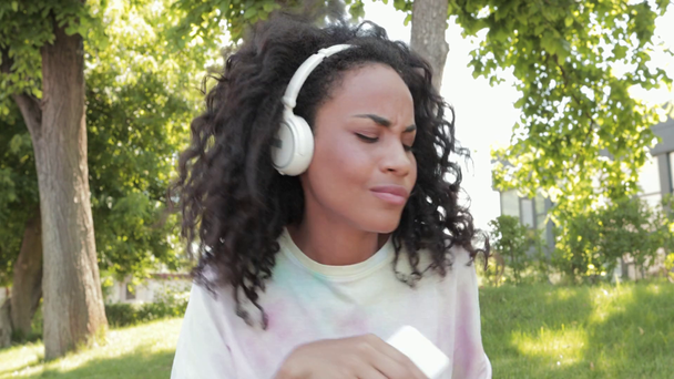 Sonriente chica afroamericana en auriculares con smartphone bailando en el parque
 - Metraje, vídeo