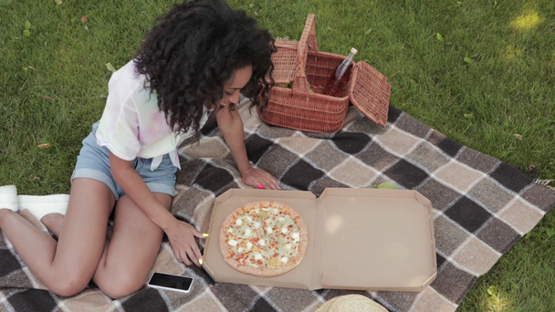 Positive Afroamerikanerin öffnet Pizzakiste auf Plaid im Park  - Filmmaterial, Video