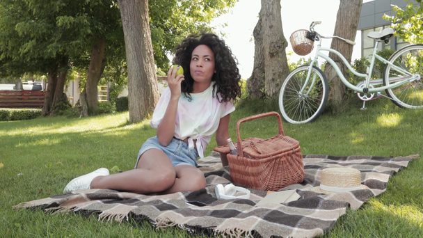 Afroamerykanka jedząca jabłko podczas pikniku na trawie w parku  - Materiał filmowy, wideo