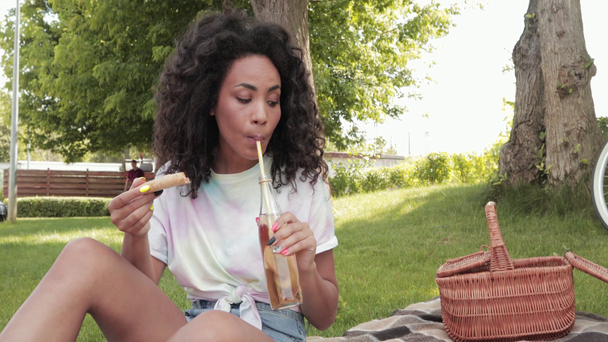 Sorridente donna africana americana che mangia pizza e beve limonata durante il picnic  - Filmati, video
