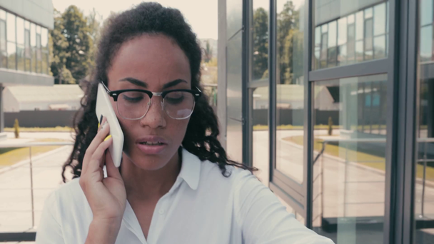 African american businesswoman talking on smartphone and walking near building - Imágenes, Vídeo