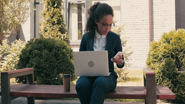Afrykański amerykański bizneswoman za pomocą laptopa i smartfona na ławce w parku  - Materiał filmowy, wideo