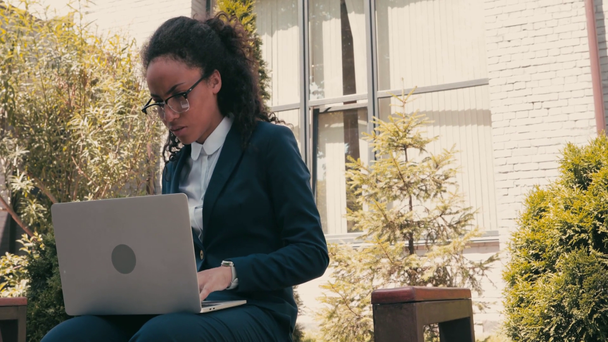 afro-americana donna d'affari bere caffè e utilizzando il computer portatile su panchina all'aperto - Filmati, video
