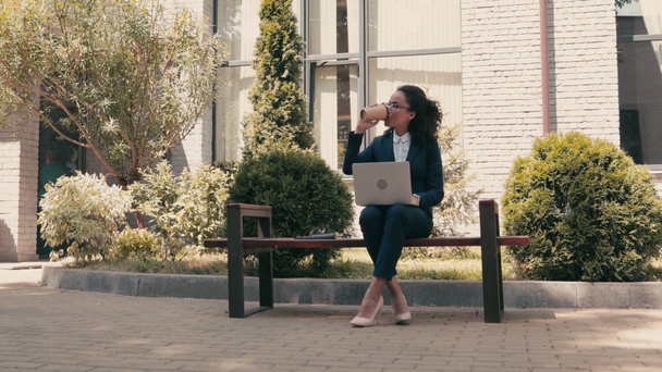 African american businesswoman with coffee and laptop on bench on urban street - Footage, Video