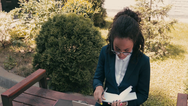 Afrikai amerikai üzletasszony jegyzetfüzettel és laptoppal a padon a városi utcában  - Felvétel, videó