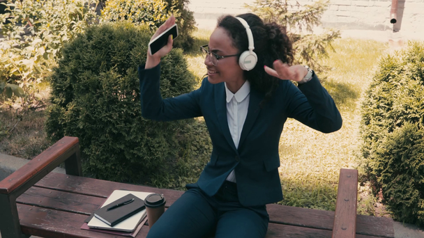 African american businesswoman in headphones dancing on bench on urban street - Materiał filmowy, wideo