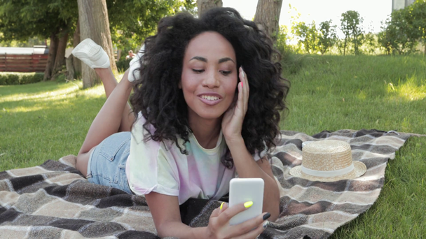 Hymyilevä afrikkalainen amerikkalainen tyttö käyttää älypuhelinta ruudullinen puistossa  - Materiaali, video