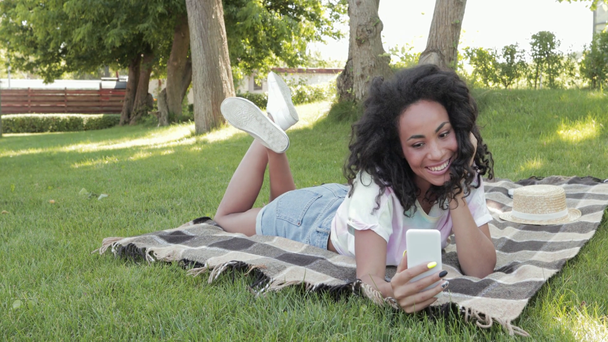 Uśmiechnięta afrykańska dziewczyna robi sobie selfie ze smartfonem podczas pikniku w parku - Materiał filmowy, wideo