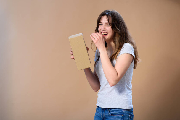 Μια διεθνής κοπέλα γελάει δυνατά με ένα κουτί στα χέρια της σε ένα απλό μπεζ φόντο. - Φωτογραφία, εικόνα