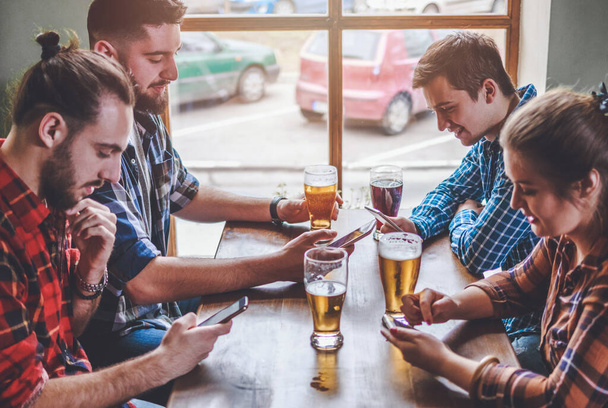 Друзья пьют пиво, занимаются серфингом на смартфонах и смеются в баре - Фото, изображение