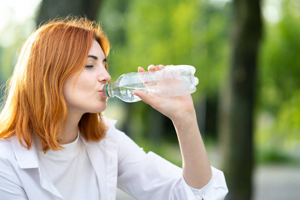Onnellinen janoinen punapää nainen juo tuoretta pullotettua vettä kesällä ulkona. - Valokuva, kuva