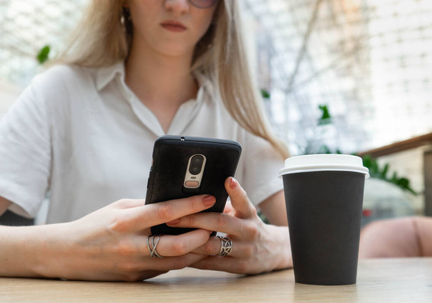 Cropped photo of cheerful young caucasian blond business woman in a white shirt typing on smartphone while resting on coffe break. Lifestyle concept. Concept of successful young women on coffe break. Nearby is a black cup of coffee. - Foto, Bild