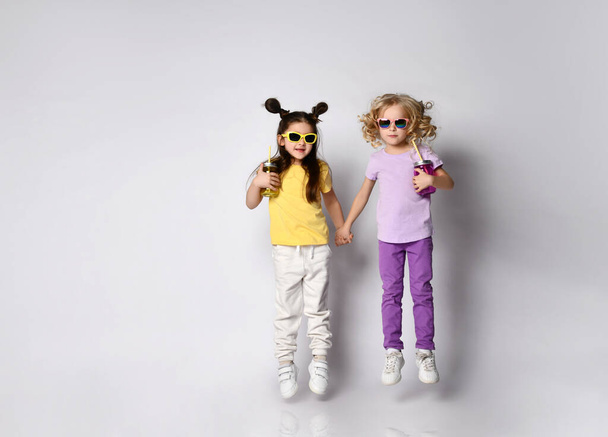 Little kids in sunglasses, colorful casual clothes. Girls holding hands and cocktail bottles, jumping up, posing isolated on white - 写真・画像