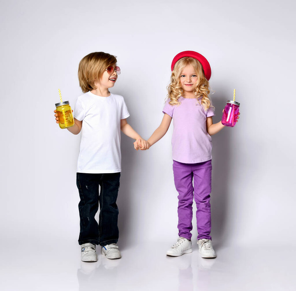 Little girl and boy in colorful casual clothes. Smiling, holding hands, yellow and pink cocktail bottles. Posing isolated on white - Zdjęcie, obraz