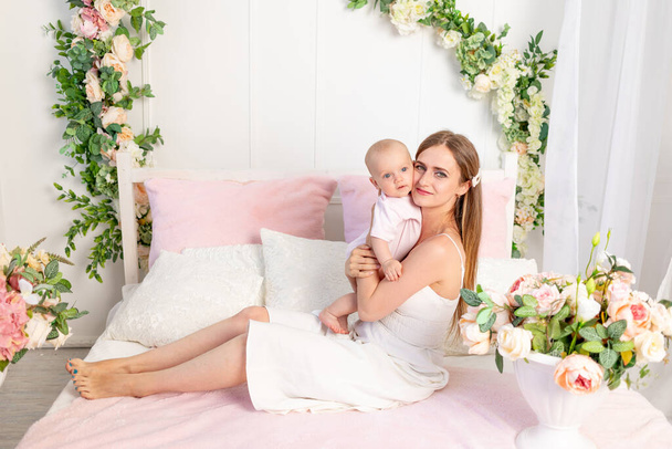 eine junge schöne Mutter mit einer 6 Monate alten Tochter im Arm sitzt auf einem weißen Blumenbeet und umarmt sie, ein Ort für Text - Foto, Bild