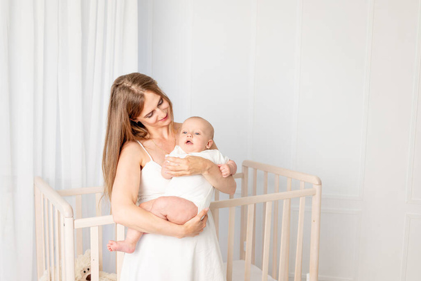 young beautiful mother holding her daughter 6 months, hugging her in the nursery standing by the crib, mother's day, place for text - Photo, Image