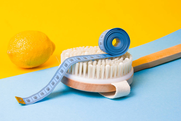 blue measuring tape on a wooden brush with natural pile of cactus and lemon on a yellow and light blue background copy space top view, anti-cellulite massage concept - Φωτογραφία, εικόνα
